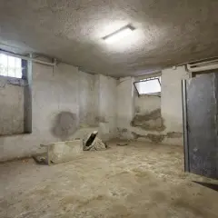 Ein leerer, feuchter Keller mit Betonwänden und -boden, bereit für die Keller sanieren. Ein Fenster mit Gitter und eine Metalltür sind sichtbar.