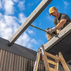 Mann misst Holz für das **carport dach erneuern kosten**. Blaue Himmel mit Wolken.