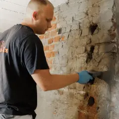 Mann mit blauen Handschuhen verputzt feuchte Kellerwand. Er arbeitet am keller trocken legen, um Feuchtigkeitsschäden zu beheben.