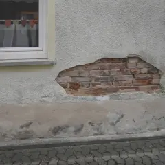 Putz löst sich im Sockelbereich, darunter Ziegelmauerwerk. Anzeichen für aufsteigende Feuchtigkeit im Sockelbereich.