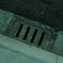 Betonierter Abfluss mit Schlitzen, daneben Asphalt. Der Abfluss erinnert an einen stinkenden revisionsschacht im keller.