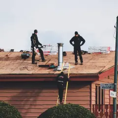 Drei Dachdecker arbeiten auf einem Hausdach. Einer bläst mit einem Laubbläser, ein anderer steht mit den Händen in den Hüften, der dritte steigt auf einer Leiter.