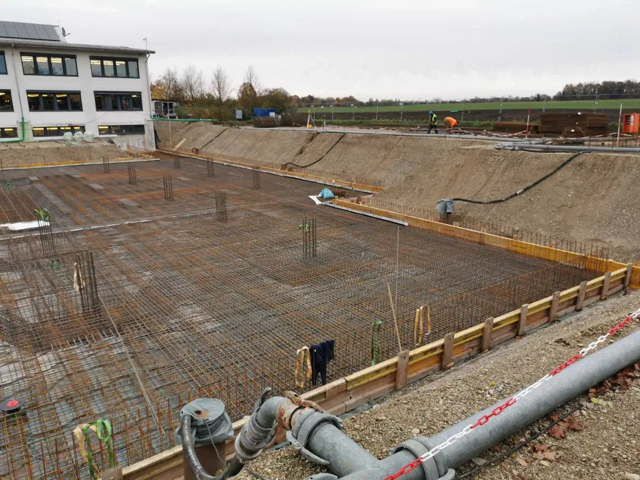 Baustelle mit Bewehrungsstahl und Schalung für eine Bodenplatte. Rohre deuten auf das grundwasser abpumpen hin, um die Arbeiten zu ermöglichen.