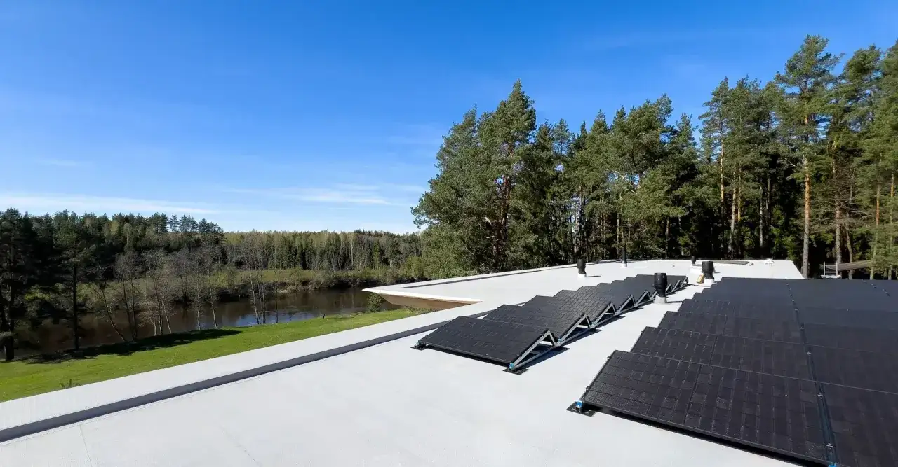 Moderne Architektur mit fpo dachbahn und Solarmodulen, umgeben von einem Fluss und einem Wald unter blauem Himmel.