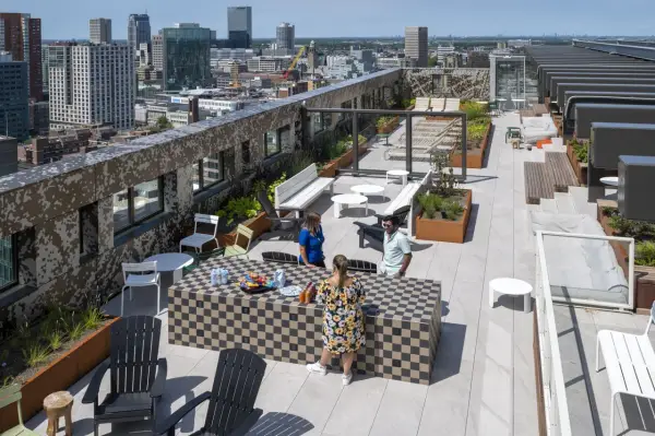 Ein begehbarer Flachdachgarten mit Stadtblick. Menschen entspannen sich auf der Dachterrasse, die mit Sitzgelegenheiten, Pflanzen und einem Schachbrett-Tisch ausgestattet ist.